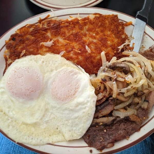 A diner breakfast with two fried eggs, hash browns, steak, and sautéed onions and mushrooms.